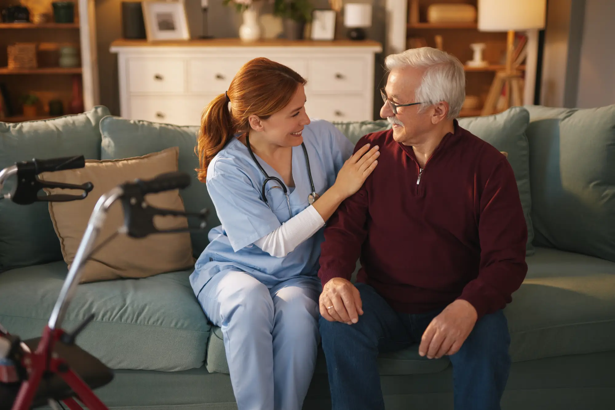 A caretaker nurse sitting next to an older gentleman on a couch with a walker in the foreground.