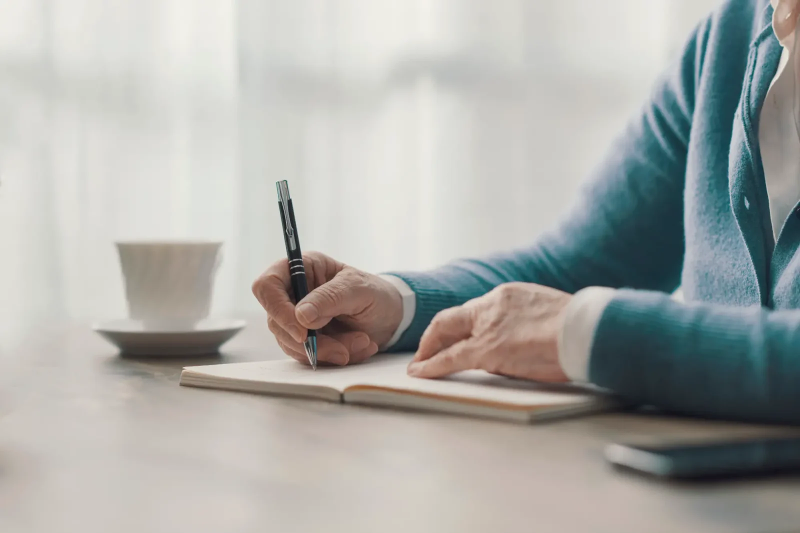 An elderly person holding a pen and writing in a notebook next to a cup of tea.