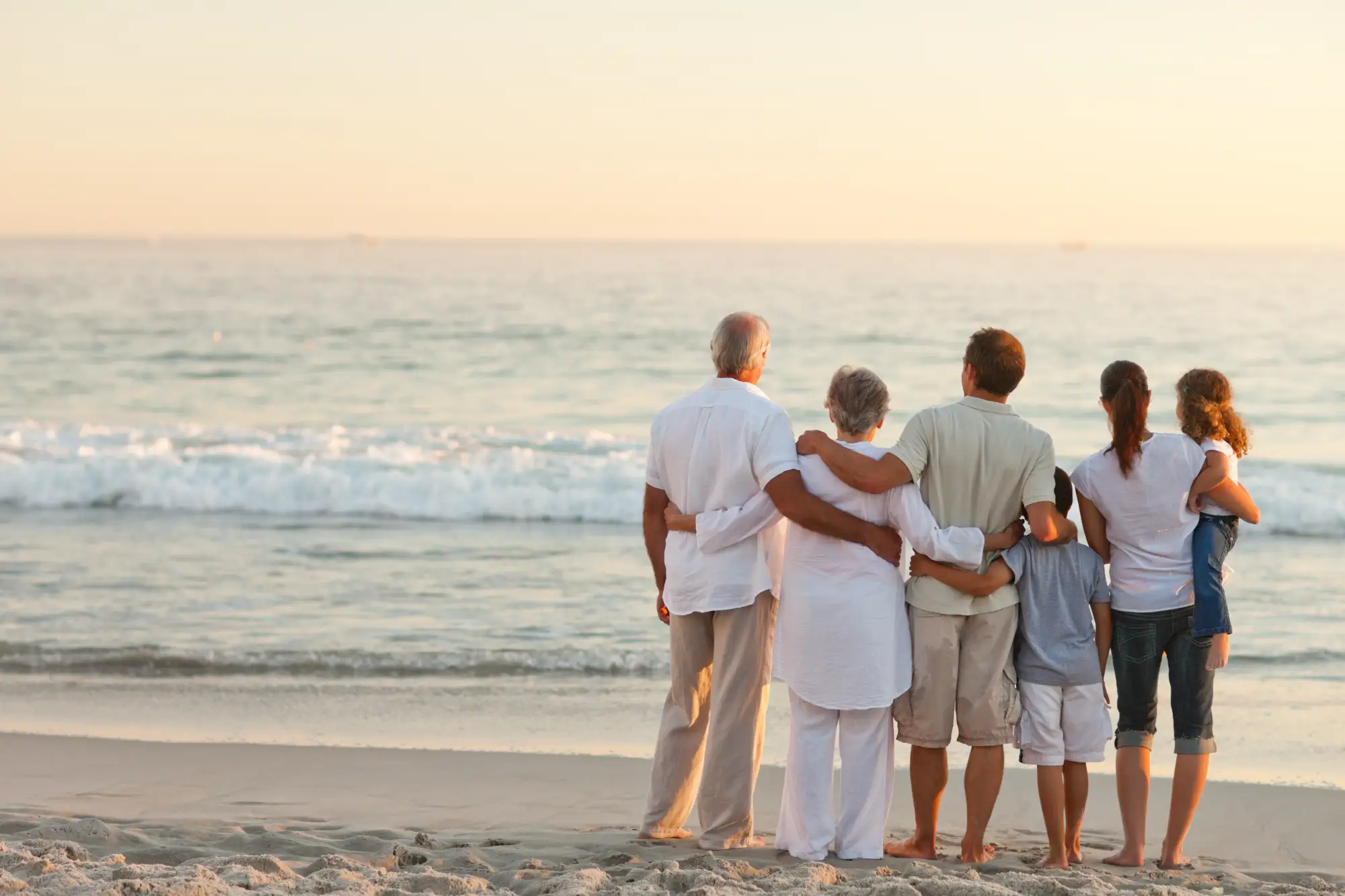 Multi-generational family of six standing on a sandy beach overlooking a vast body of water.