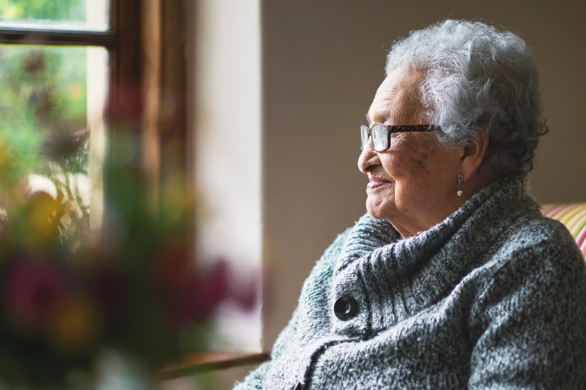 An elderly woman with glass smirking while looking out a window.