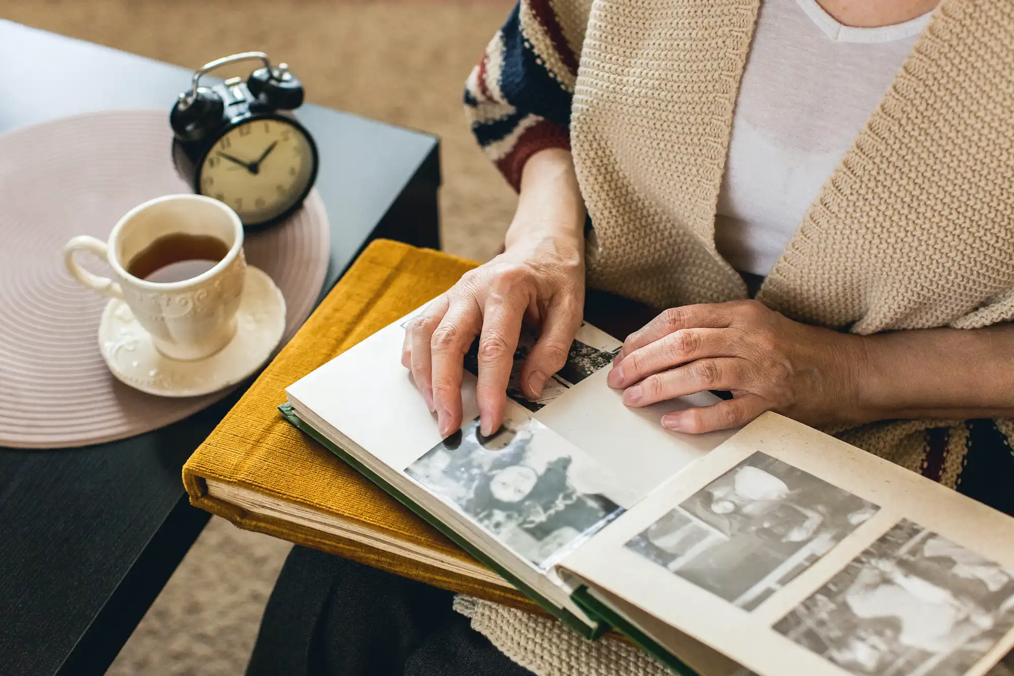 An elderly person looking at old photo albums sitting in her lap next to an end table with a cup of tea and old alarm clock.