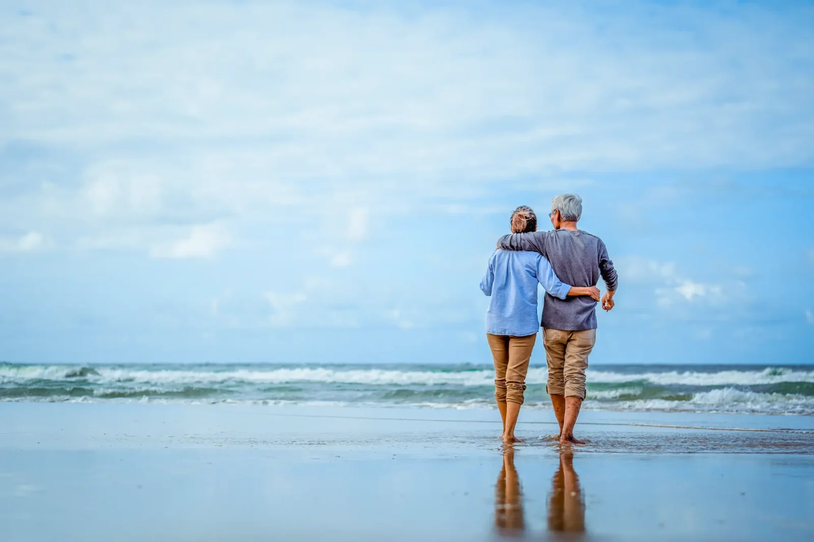 An elderly couple with their arms wrapped around each other standing in very shallow water on a beach overlooking a large body of water.